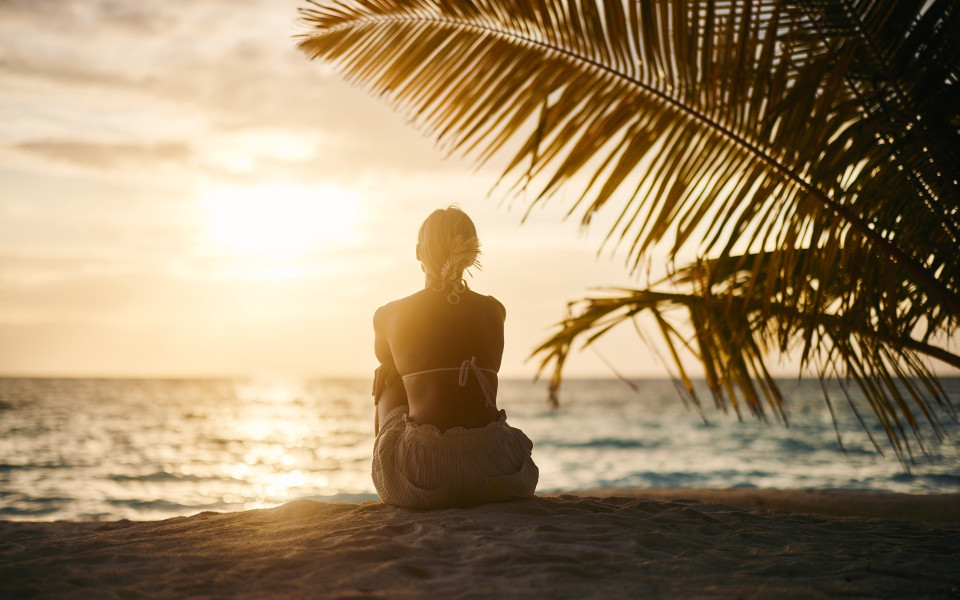 Frau sitzt am Strand unter Palmen bei Sonnenuntergang