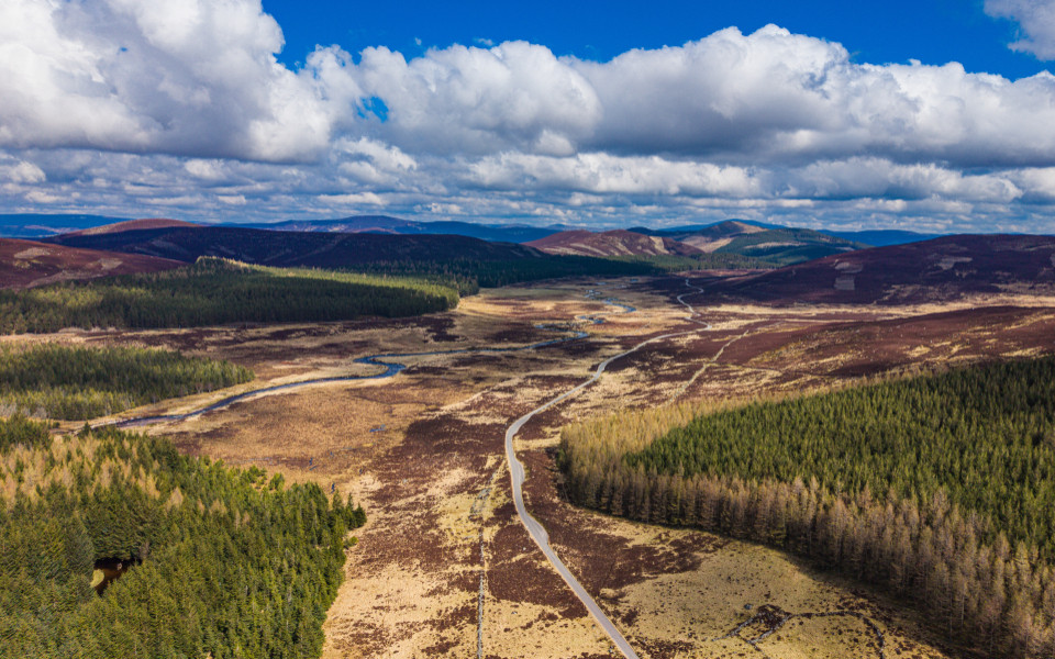 es ist Aberdeenshire in Schottland  11:16 Luftaufnahme der Aberdeenshire-Landschaft in Schottland mit gewundenem Fluss, schmaler Stra&szlig;e durch Heideland, Nadelw&auml;ldern und Bergh&uuml;geln unter blauem Himmel mit Wolken.